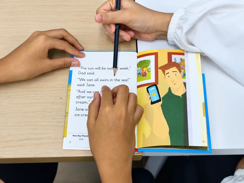 Pandangan dekat guru menggunakan pensel untuk membimbing pelajar mengikuti perkataan dalam buku bacaan.